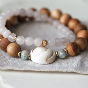 Può includere: Due braccialetti di perline. Un braccialetto presenta perline di quarzo rosa chiaro e dettagli dorati. L'altro ha perline di legno e un accento di conchiglia bianca. I braccialetti poggiano su una superficie di lino grigio.