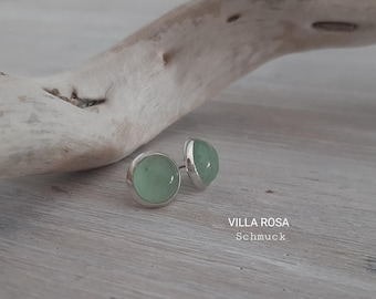 May include: A pair of silver stud earrings with round, light green stones. The earrings are on a light wood surface. The text "VILLA ROSA Schmuck" is visible below the earrings.