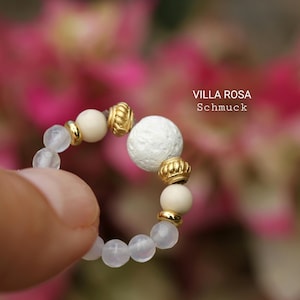 Può includere: Un anello di perline bianche e dorate con una grande perla bianca al centro. L'anello è su un dito contro uno sfondo floreale rosa e bianco. Il testo "VILLA ROSA Schmuck" è visibile nell'immagine.