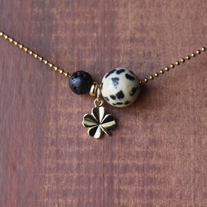Peut inclure: Un collier à chaîne dorée avec une perle de lave noire, une perle blanche et noire tachetée et un charm en forme de trèfle à quatre feuilles doré. Le collier est présenté sur une surface en bois brun.
