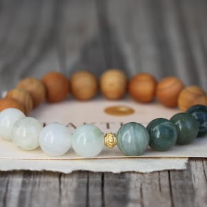 Könnte beinhalten: Ein Armband mit einer Mischung aus hellgrünen, weißen und braunen Holzperlen. Das Armband hat eine goldfarbene Perle. Das Armband wird auf einem Stück Papier mit dem Aufdruck "NAKTI" präsentiert.