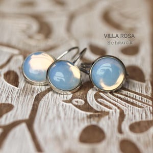 Pode incluir: Um par de brincos de prata com pedras de opala azul claro. Os brincos estão sobre uma superfície de madeira com um design entalhado. O texto "VILLA ROSA Schmuck" é visível na imagem.