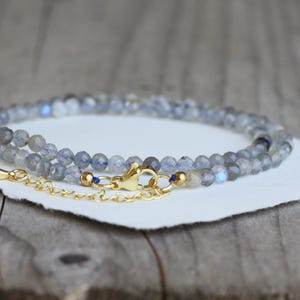 Peut inclure: Un collier délicat composé de petites perles de labradorite grise translucide à facettes avec des reflets bleus irisés. Le collier est doté d'un fermoir mousqueton doré poli et d'une courte chaînette d'extension dorée.