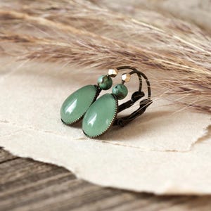 Op de afbeelding: Oorhangers met ovale, lichtgroene edelstenen en een donker metalen frame. Elke oorbel heeft een kleine, ronde, groene kraal en een goudkleurige accentkraal. De achtergrond bevat gedroogde grassen en getextureerd papier.