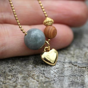 Könnte beinhalten: Eine goldfarbene Halskette mit einem kleinen Herzanhänger. Die Halskette hat eine goldfarbene Kette, eine graue Perle, eine Holzperle und ein dekoratives Goldelement. Der Herzanhänger ist glatt und glänzend.