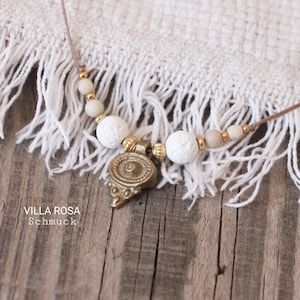 Puede incluir: Un collar con un colgante y cuentas. El colgante es de color bronce con detalles intrincados. El collar tiene cuentas blancas, beige y doradas. El cordón es de color marrón claro. El texto "VILLA ROSA Schmuck" es visible.