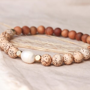May include: A stretch bracelet featuring a combination of round brown wooden beads, round beige beads with dark brown speckles, two small gold-toned metal spacer beads, and a single central white oval-shaped pearl bead.