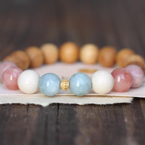 Könnte beinhalten: Armband mit Holz-, rosa-, weißen und hellblauen Edelsteinperlen. Das Armband ist auf einem elastischen Band aufgefädelt und hat eine goldfarbene Abstandshülse. Das Armband wird auf einem Stück Papier präsentiert. Die Perlen haben einen Durchmesser von ca. 1 cm.