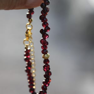 May include: A beaded necklace featuring deep red gemstones and gold-coloured metal accents. The necklace is held up, showcasing the rich colour of the beads and the intricate design of the clasp and chain. The gemstones appear to be approximately 0.6 cm in diameter.