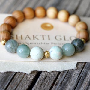 Pode incluir: Uma pulseira de contas com contas castanhas claras e verde-azuladas claras alternadas. A pulseira é exibida em um cartão creme com o texto "SHAKTI GLO" e "handgemachter Perlen". A pulseira está em uma superfície de madeira.