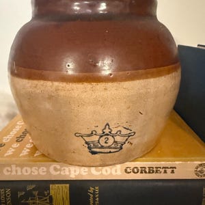 Op de afbeelding: Een tweekleurige stenen pot met een donkerbruin geglazuurd bovengedeelte en een lichtbeige ondergedeelte. De voorzijde is voorzien van een gestempeld blauw kroonicoon met het cijfer 2 erin. De pot bevat een bijpassend deksel met een knop.