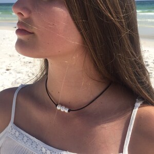 May include: A simple black choker necklace with three white pearls. The necklace is worn by a person with long brown hair.