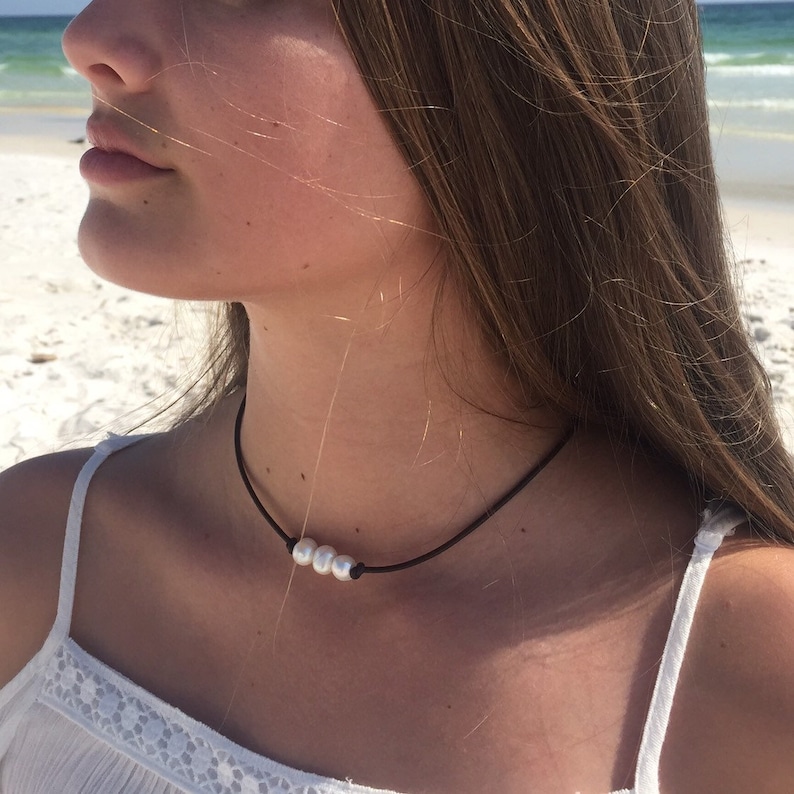 May include: A simple black choker necklace with three white pearls. The necklace is worn by a person with long brown hair.