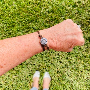 May include: A brown leather bracelet with a silver daisy flower charm. The bracelet is tied with knots and is worn on a wrist. The background is green grass.