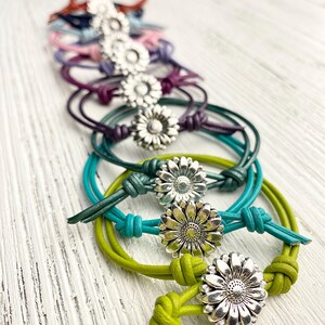 May include: A collection of daisy-adorned bracelets in various colors, including green, teal, purple, and pink. Each bracelet features a silver-toned daisy charm and knotted leather cord, creating a charming and handcrafted look. The bracelets are arranged on a white wooden surface.