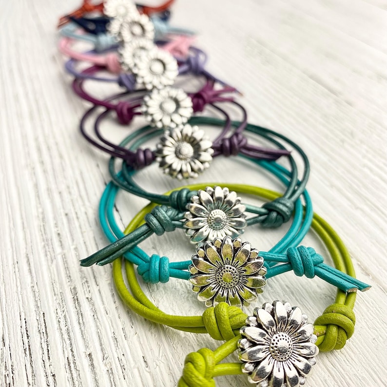 May include: A collection of daisy-adorned bracelets in various colors, including green, teal, purple, and pink. Each bracelet features a silver-toned daisy charm and knotted leather cord, creating a charming and handcrafted look. The bracelets are arranged on a white wooden surface.
