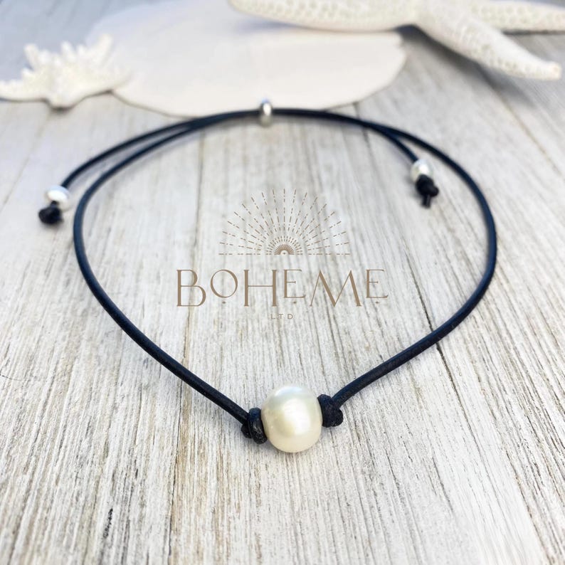 Puede incluir: Un collar de cord&oacute;n negro con una sola perla blanca luminosa. El collar tiene nudos ajustables y detalles plateados. La palabra "BOHEME" est&aacute; impresa en el fondo.