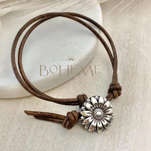 May include: A brown leather bracelet with a silver daisy flower charm. The bracelet features a double-strand design and is knotted at the ends. The word "BOHEME" is visible in the background.