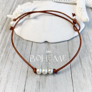 May include: A brown leather choker necklace with three white pearls. The necklace is tied with a knot at the back.