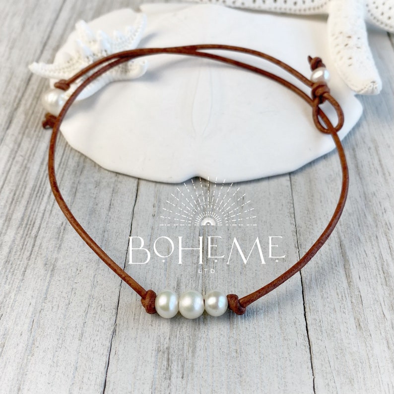 May include: A brown leather choker necklace with three white pearls. The necklace is tied with a knot at the back.