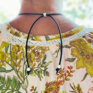 Puede incluir: Collar gargantilla negro con un dise&ntilde;o delicado. El collar presenta un fino cord&oacute;n negro con un peque&ntilde;o acento de perla en la parte delantera y lazos ajustables. El collar se usa sobre una blusa con estampado floral.