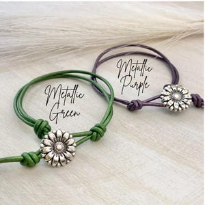 May include: Two adjustable bracelets, one metallic green and one metallic purple, each featuring a daisy-shaped charm. The bracelets are displayed on a light-colored surface, with the words "Metallic Green" and "Metallic Purple" written on them.
