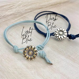 May include: Two adjustable bracelets with daisy flower charms. One bracelet is light blue, the other is dark blue. The text "Light Blue" and "Dark Blue" are written on the image. The bracelets are on a white wooden surface.