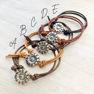 May include: Five leather bracelets with silver daisy flower accents. The bracelets are in various shades of brown, from light tan to dark brown and black. Each bracelet features a knotted closure.