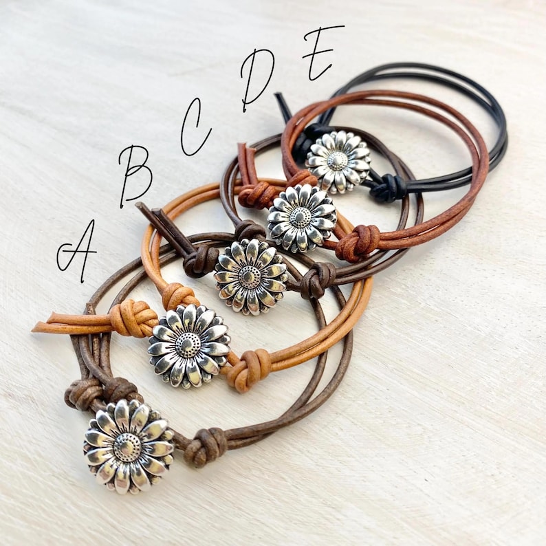 May include: Five leather bracelets with silver daisy flower accents. The bracelets are in various shades of brown, from light tan to dark brown and black. Each bracelet features a knotted closure.