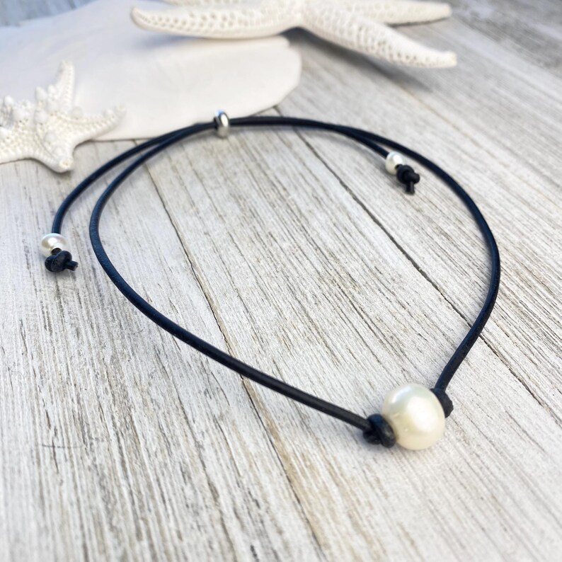 Puede incluir: Tobillera de cord&oacute;n negro con una perla blanca y redonda grande. El cord&oacute;n est&aacute; anudado a ambos lados de la perla y en los extremos, con una peque&ntilde;a cuenta plateada cerca del cierre. Fondo con estrellas de mar blancas.