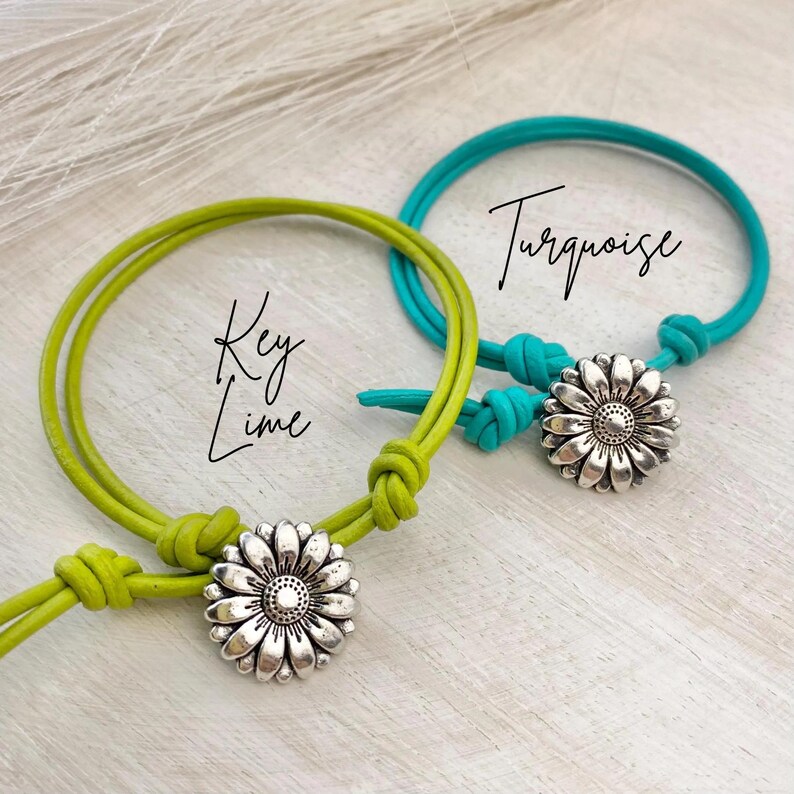 May include: Two bracelets with daisy charms. One is key lime green, the other turquoise. Each bracelet has a silver-colored daisy charm and is labeled with the color name in cursive script. The bracelets are on a light wood-grain surface.