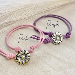 May include: Two bracelets, one pink and one purple, each with a silver daisy-shaped charm. The bracelets have a knotted design and the word "Pink" and "Purple" are written on the image.