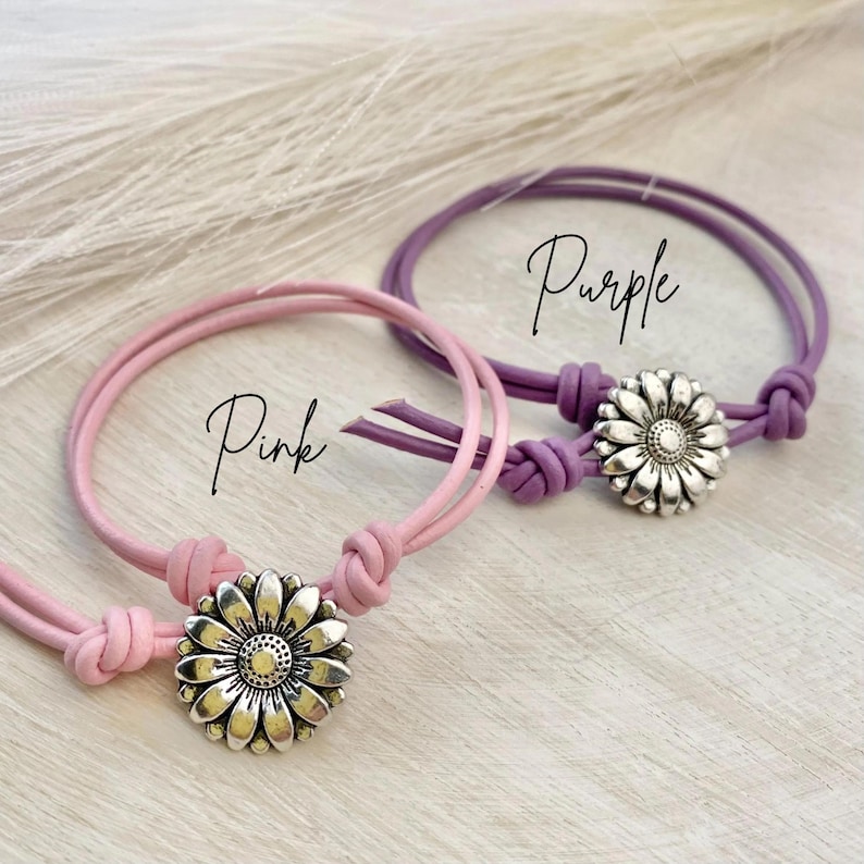 May include: Two bracelets, one pink and one purple, each with a silver daisy-shaped charm. The bracelets have a knotted design and the word "Pink" and "Purple" are written on the image.