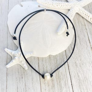 Puede incluir: Un collar gargantilla de cuero negro con un solo colgante de perla blanca. El collar tiene un dise&ntilde;o delicado con nudos ajustables y un cierre plateado. El fondo presenta estrellas de mar blancas y un d&oacute;lar de arena blanco sobre una superficie de madera clara.