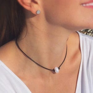 Puede incluir: Un collar minimalista con un solo colgante de perla blanca luminosa sobre un fino cord&oacute;n marr&oacute;n oscuro. El collar se combina con un peque&ntilde;o pendiente de bot&oacute;n plateado y redondo. La joya se lleva sobre una camisa blanca.