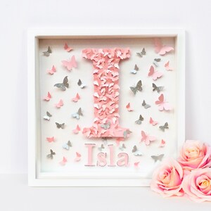 May include: A white frame with a pink letter I made of butterflies and the name Isla written below. The frame is surrounded by pink and silver butterflies.