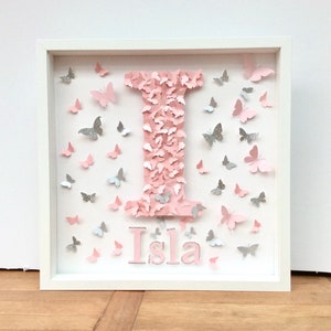 May include: A white frame with a pink letter I made of pink butterflies and the name Isla written below in pink letters. The background is filled with pink and silver butterflies.
