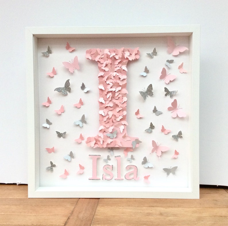 May include: A white frame with a pink letter I made of pink butterflies and the name Isla written below in pink letters. The background is filled with pink and silver butterflies.
