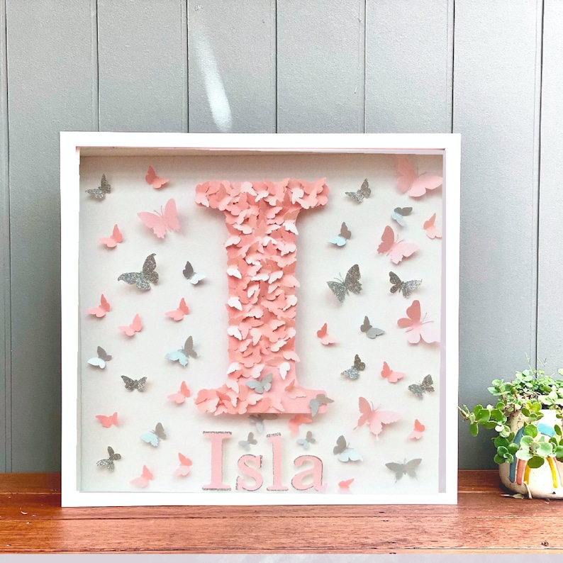 May include: A white frame with a pink letter I made of butterflies. The name Isla is written below the letter. The frame is filled with pink and silver butterflies.
