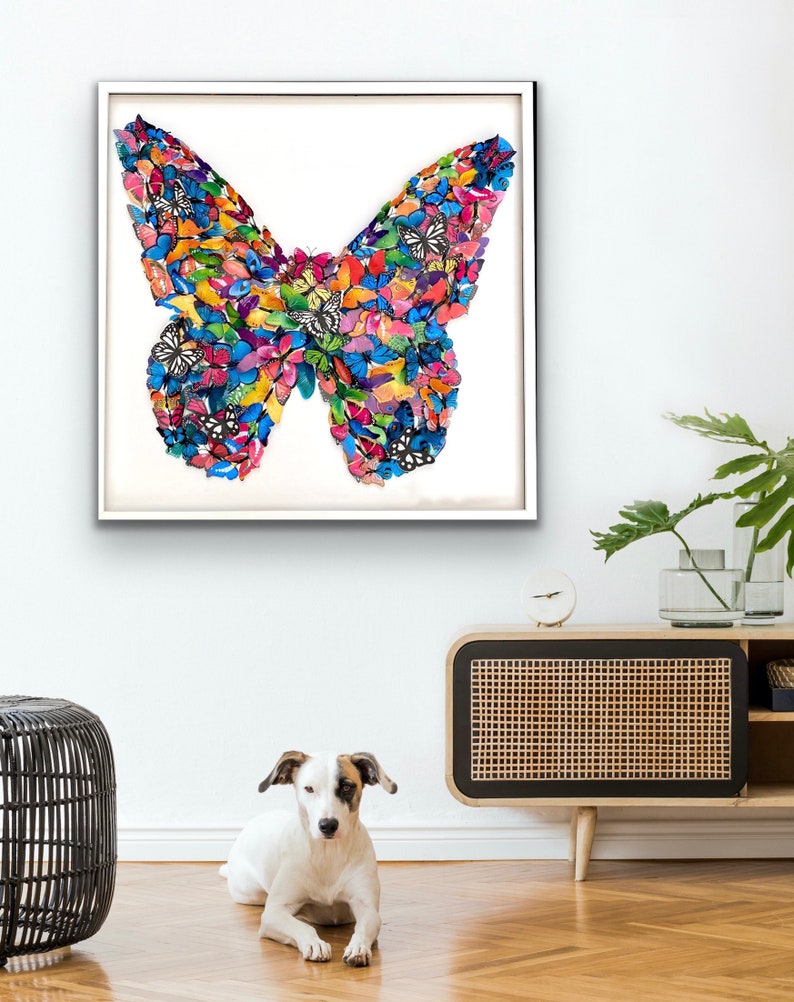 May include: A framed artwork featuring a colorful butterfly made of smaller butterflies. The butterfly is a collage of various colors and patterns, including blue, green, yellow, orange, and pink. The artwork is displayed on a white wall in a modern living room setting.