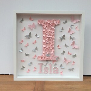 May include: A white frame with a pink letter I made of butterflies. The letter is surrounded by pink and silver butterflies. The name Isla is written below the letter in pink.