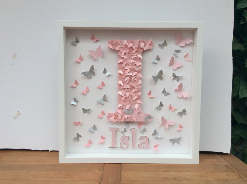 May include: A white frame with a pink letter I made of butterflies. The letter is surrounded by pink and silver butterflies. The name Isla is written below the letter in pink.