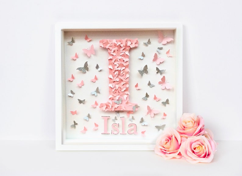 May include: A white frame with a pink letter I made of butterflies and the name Isla written below. The frame is surrounded by pink and silver butterflies.