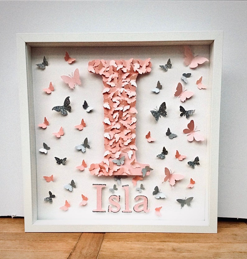 May include: A white picture frame with a pink letter I made of butterflies. The name Isla is written below the letter in pink.