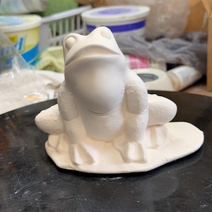 May include: A ceramic frog figurine in an off-white color. The frog is sitting on a lily pad base. The figurine is unpainted and ready for customization. The frog has large eyes and detailed features.
