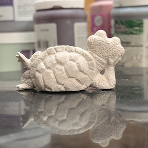 May include: An unpainted ceramic turtle figurine in a reclining pose, resting its head on one front flipper. The sculpture features a detailed, textured shell with a geometric pattern and a rough, matte off-white surface finish.