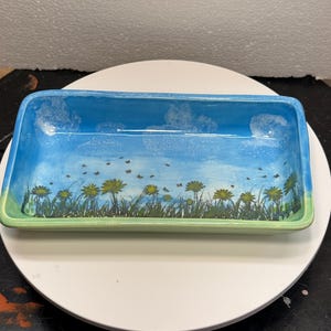 May include: A rectangular ceramic tray featuring a hand-painted landscape scene. The interior displays a bright blue sky with white clouds above a field of green grass and yellow dandelion flowers. The exterior rim is a soft, light green color.