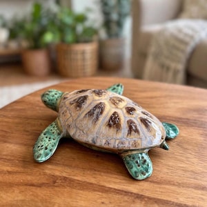 May include: A ceramic sea turtle figurine featuring a glossy, multi-toned shell with shades of tan, cream, and dark brown. The head and flippers are a textured, mottled teal green with dark spots, showcasing a smooth, glazed finish throughout.