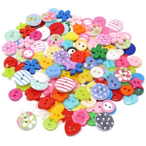 May include: A large collection of colourful plastic buttons in various shapes, including stars, hearts, flowers, and circles. The buttons are arranged in a pile on a white background.