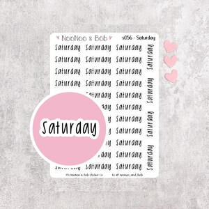 Könnte beinhalten: Ein Aufkleberbogen mit mehreren Zeilen des Wortes Saturday in einer schwarzen Handschrift-Schriftart. Ein großer runder rosa Aufkleber mit dem Wort Saturday in Weiß liegt auf dem Bogen. Drei kleine rosa Herzaufkleber sind enthalten.